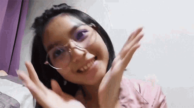 a young girl wearing glasses and a pink shirt is smiling and waving at the camera .