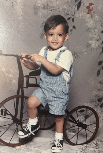 a young boy sitting on a tricycle wearing blue overalls