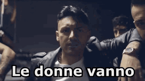 a man in a leather jacket is standing in front of a group of men with the words le donne vanno written above him .