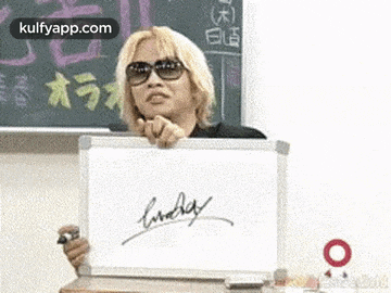 a man is holding a white board with a signature on it in front of a blackboard .