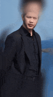 a man in a suit and black shirt is standing in front of a body of water