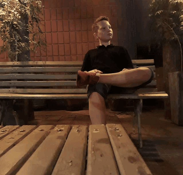 a young boy sits on a wooden bench with his feet crossed