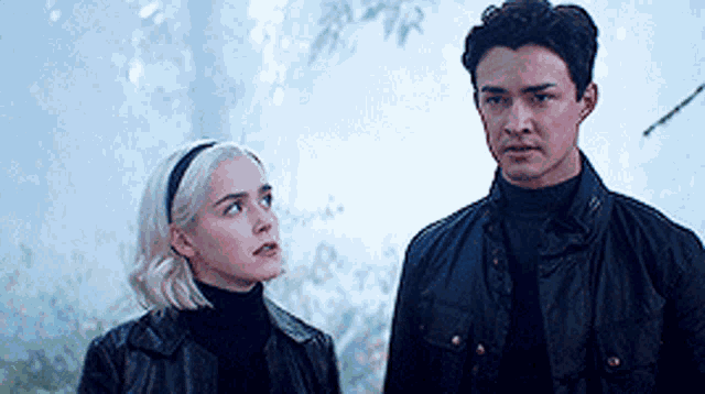 a man and a woman are standing next to each other in a forest looking at each other .