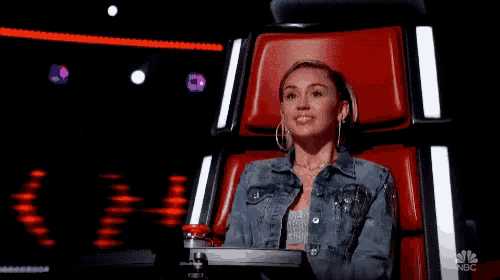 a woman in a denim jacket is sitting in a red chair with her hands folded and smiling .