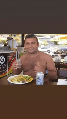 a shirtless man sits at a table with a plate of food and a can of fanta soda