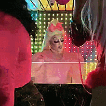 a drag queen with a pink bow on her head is standing in front of a mirror