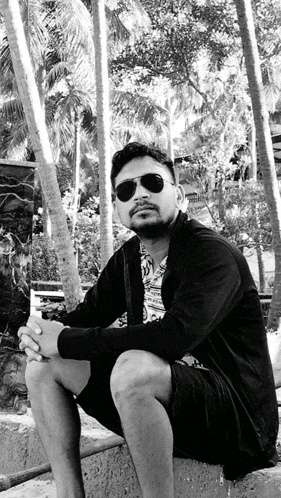 a black and white photo of a man wearing sunglasses sitting on a ledge