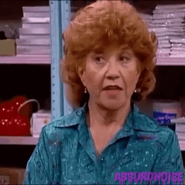 a woman with red hair and a blue shirt is standing in front of a shelf with papers on it .