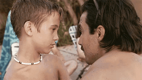 a man and a boy are looking at each other on a beach . the boy is wearing a necklace .