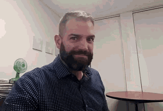 a man with a beard is taking a selfie in a room with a table and a fan .