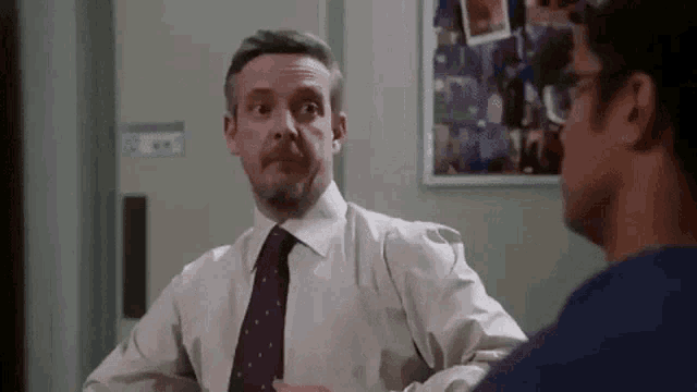 a man in a white shirt and purple tie is talking to another man in a room .