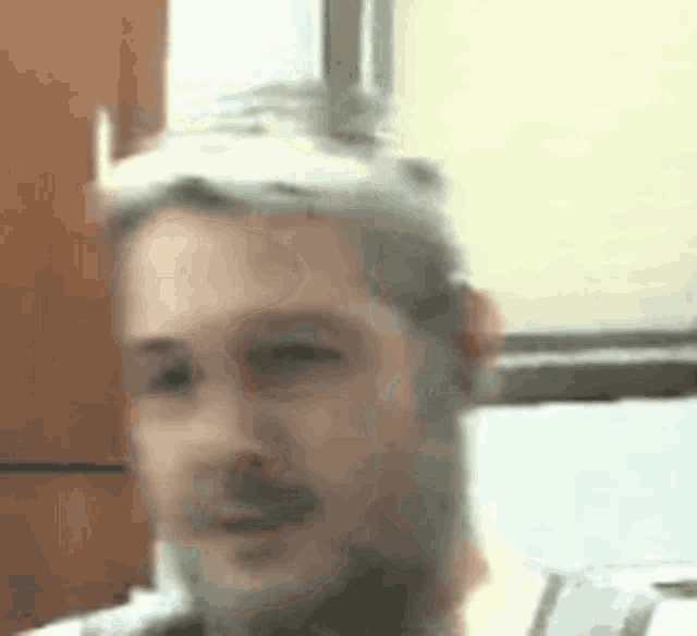 a man with a beard is sitting in front of a window and making a funny face .