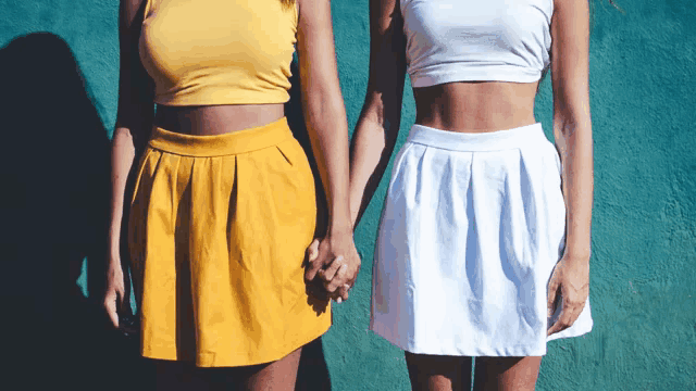 two women wearing skirts and crop tops hold hands