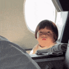a little girl looking out of an airplane window with a newspaper laying on the seat next to her