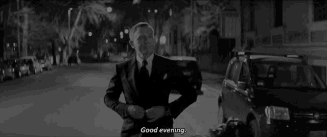 a man in a suit and tie is standing on a street and says good evening