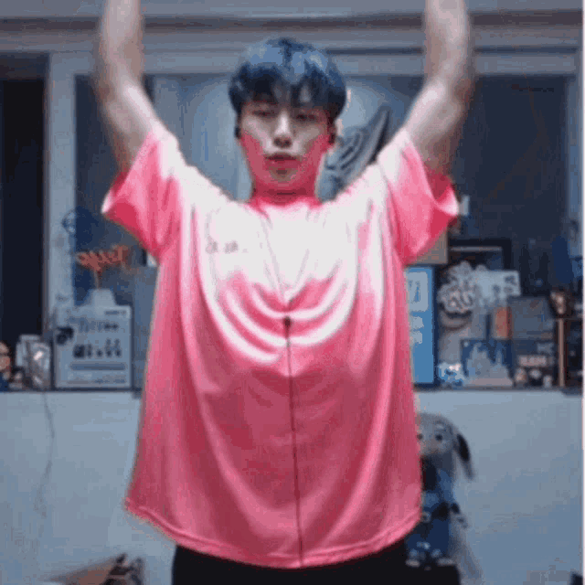 a young man with blue hair is wearing a pink shirt and headphones and raising his arms in the air .