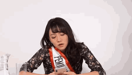a woman wearing a sash with the word pemi on it is sitting at a table looking at her phone .