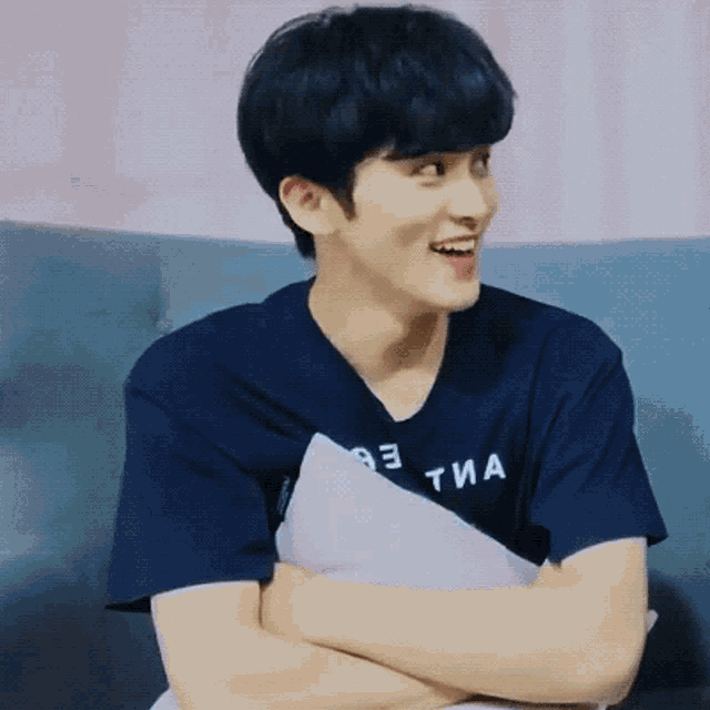 a young man in a blue shirt is sitting on a couch with his arms crossed and a pillow .