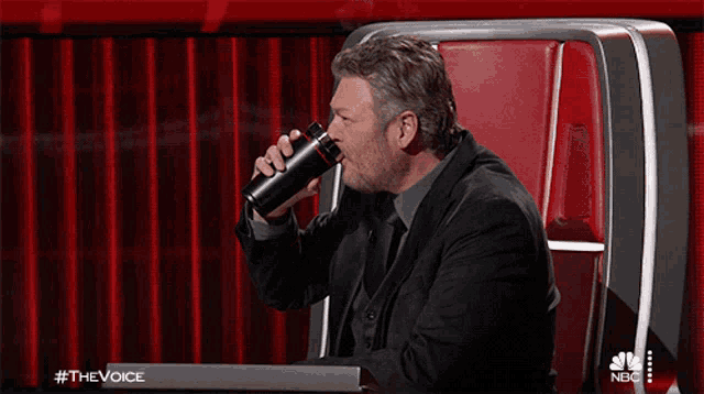 a man sitting in a chair drinking from a black cup with #thevoice on the bottom right