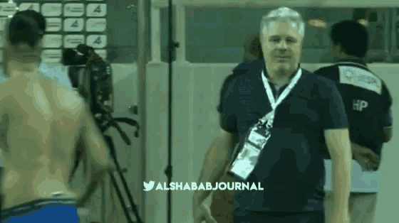 a man with a lanyard around his neck stands in front of a camera with the twitter hashtag alshababjournal at the bottom