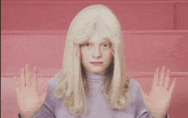 a woman wearing a white wig and a purple turtleneck shows off her hands