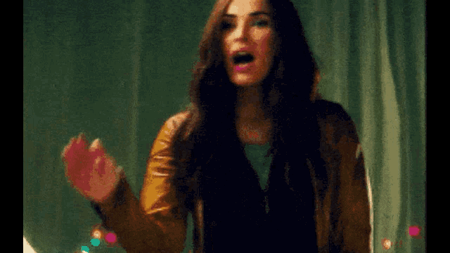 a woman in a brown leather jacket is waving her hand in front of a green curtain .