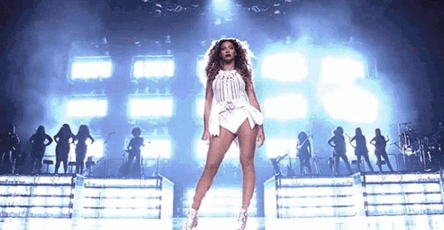 a woman in a white dress is standing on a stage in front of a group of dancers .