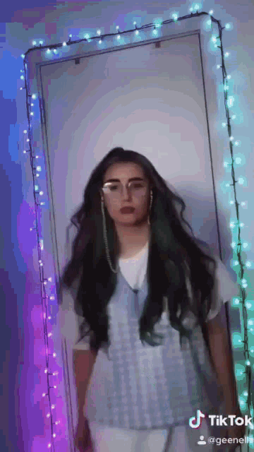 a woman with long hair is standing in front of a mirror with christmas lights hanging on the wall behind her .