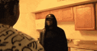 a man in a hooded jacket is standing in a kitchen with another man .
