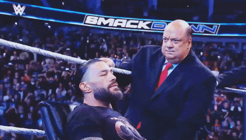 a man in a suit and tie stands next to a man in a chair in a wrestling ring