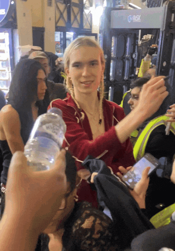 a woman in a red jacket is surrounded by people holding a bottle of water and a cell phone