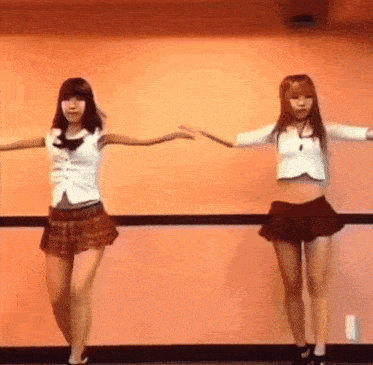 two girls in school uniforms are dancing in front of an orange wall .