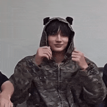a young man wearing a camouflage jacket and a hoodie with bear ears on it .