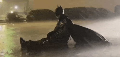 a man in a batman costume is sitting on top of another man in a puddle of water .