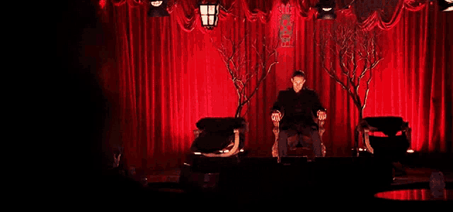 a man is sitting in a chair in front of a red curtain with a lantern hanging from the ceiling
