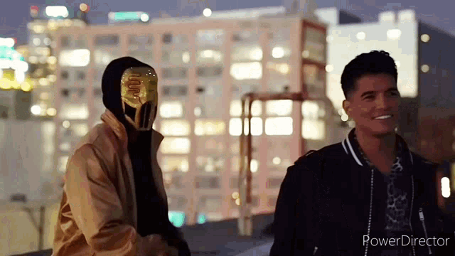 two men are standing next to each other in front of a city at night . one of the men is wearing a mask .