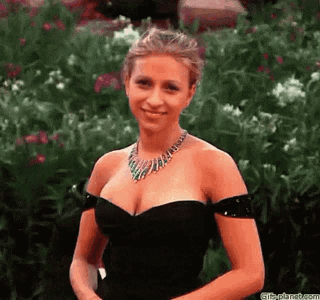 a woman wearing a black dress and a necklace is standing in front of a bush .