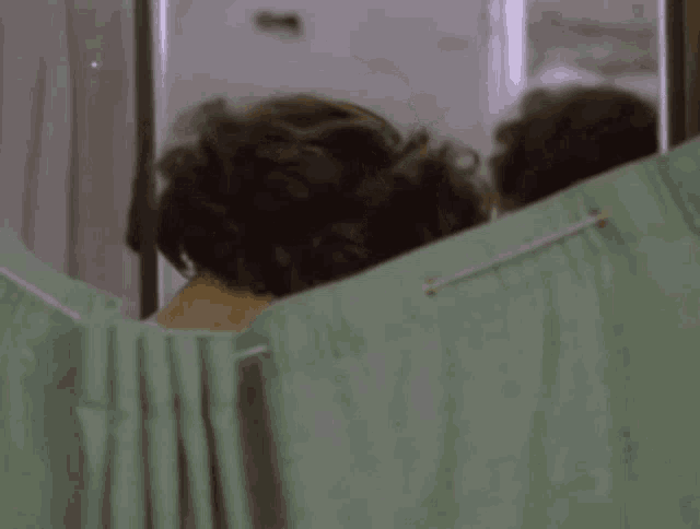 a woman is looking at herself in a mirror behind a green curtain in a bathroom .