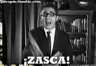 a man in a suit and tie is standing in front of a bookshelf and shouting zasca .