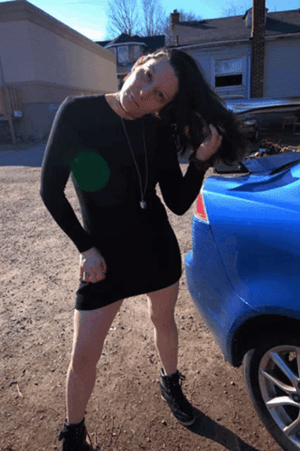 a woman in a black dress stands in front of a blue car