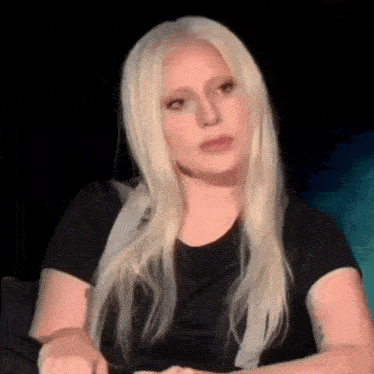 a woman with blonde hair is wearing a black shirt and sitting in a dark room .