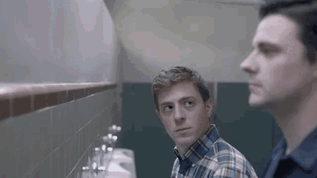 two men are standing next to each other in a public restroom looking at each other .