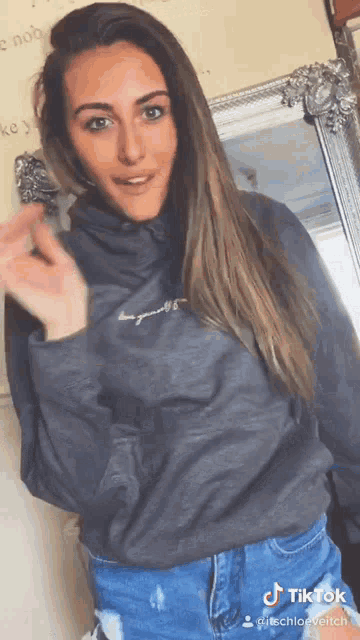 a woman wearing a grey sweatshirt and blue ripped jeans has a tiktok sticker on her shoulder