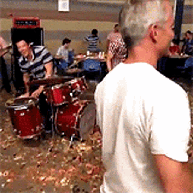 a man in a white shirt is standing in front of a drum set .