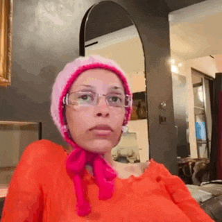 a woman wearing a pink hat and glasses is sitting in front of a mirror in a room .