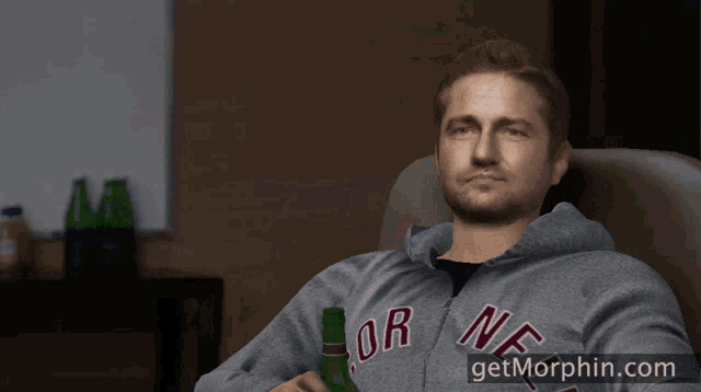 a man is sitting in a chair holding a bottle of beer and wearing a grey hoodie that says or on it