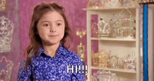 a little girl in a blue shirt is standing in front of a shelf of trophies and tiaras .