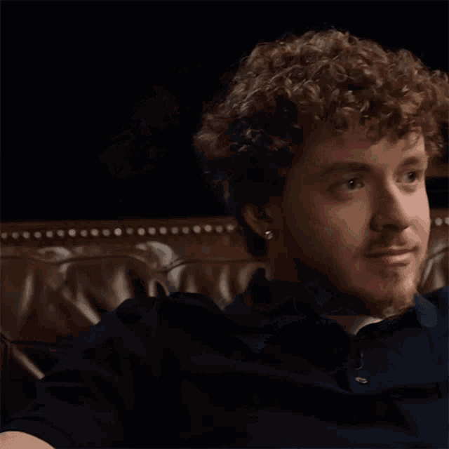 a man with curly hair and a beard is sitting on a couch looking at the camera .