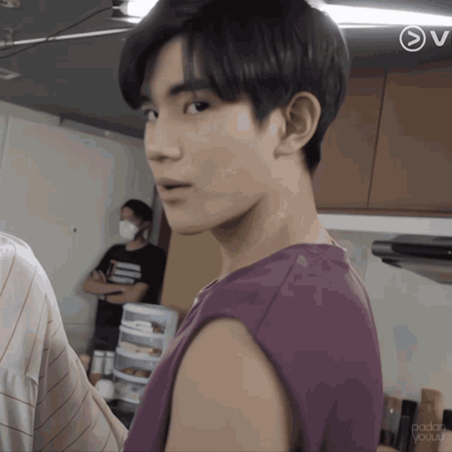 a man in a purple shirt is standing in a kitchen with a man wearing a mask behind him