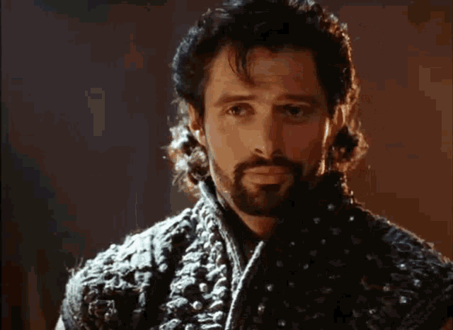 a man with a beard and long hair is wearing a black vest with studs on it .